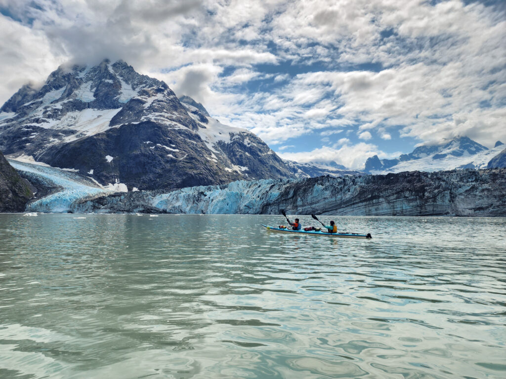 A kayak on a serene glacial lake with a young couple seated and paddling together. A glacial mountain in the background with blue sky and clouds.