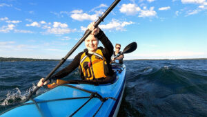 A young woman and her husband kayaking in an open body of water on a sunny day. They are joyfully pushing through the water with their paddles, enjoying themselves tremendously.