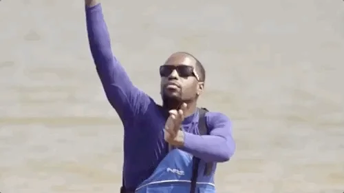 A man on a sit-on kayak, wearing sunglasses, raises his hands in a flowing motion while looking upwards, as if doing tai chi; a moment of tranquility.